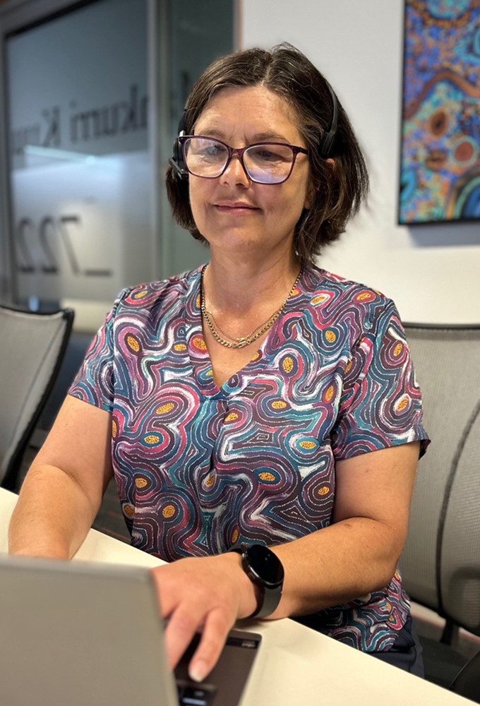 Sandra, a female ATO Evergeen Program graduate, smiling, seated at a desk working on a laptop in an ATO office, wearing an Indigenous print blouse, with Indigenous artwork on the wall.