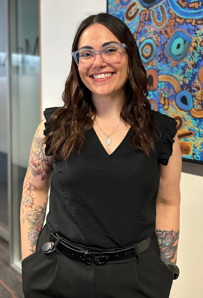 Ayla, a young female Evergreen Program graduate, standing smiling in an ATO office in front of an Indigenous artwork on the wall, wearing a black top and trousers.